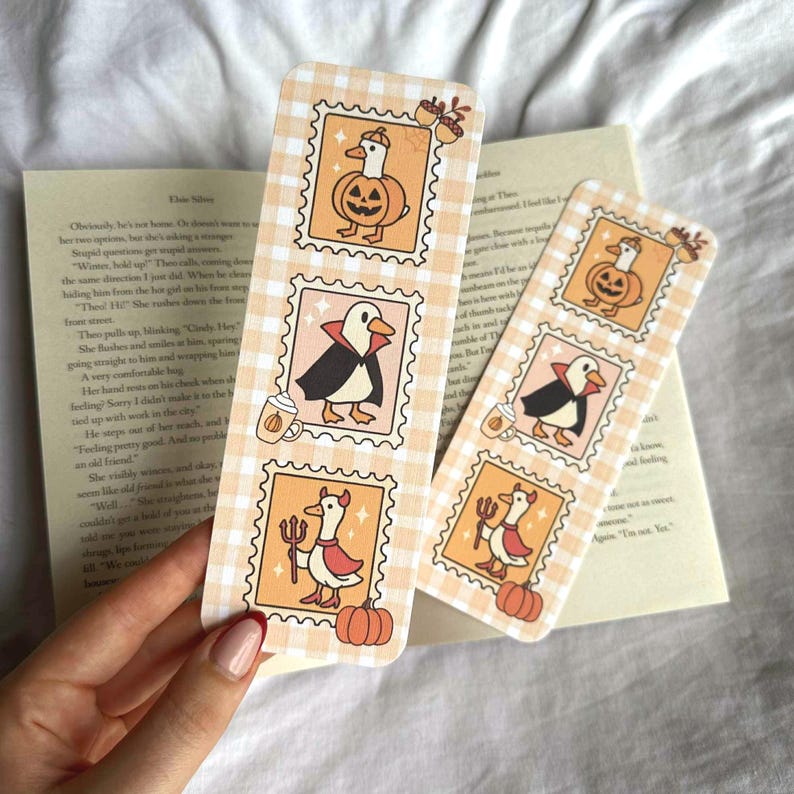 Op de afbeelding: Twee Halloween-thema bladwijzers met een lichtoranje ruitpatroon. Elke bladwijzer heeft drie postzegel-stijl illustraties van eenden in kostuums, waaronder een pompoen, vampier en duivel. Een boek staat op de achtergrond.