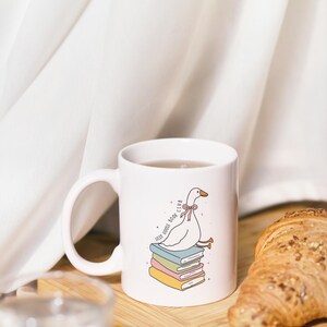 May include: White ceramic mug featuring a cartoon goose sitting on a stack of books. The mug has the text "Jolly Goose Book Club" and is filled with a beverage. A croissant sits on a wooden board next to the mug.