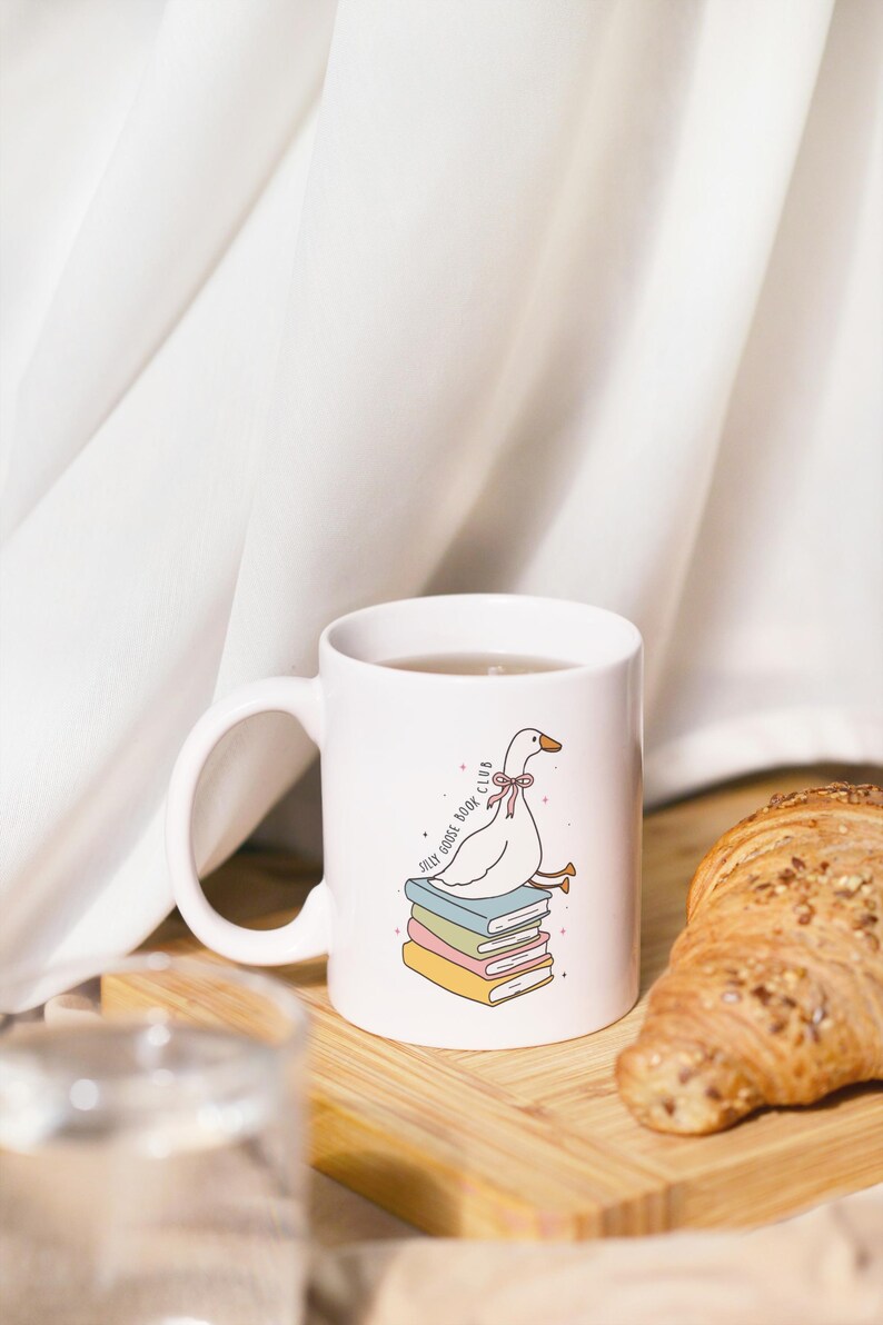 May include: White ceramic mug featuring a cartoon goose sitting on a stack of books. The mug has the text "Jolly Goose Book Club" and is filled with a beverage. A croissant sits on a wooden board next to the mug.
