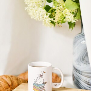 May include: White ceramic mug with a cartoon goose wearing a bow, perched on a stack of books. The mug has the text "Self Goose Book Club". A spoon and coffee beans sit on a wooden cutting board, with a croissant and flowers in the background.