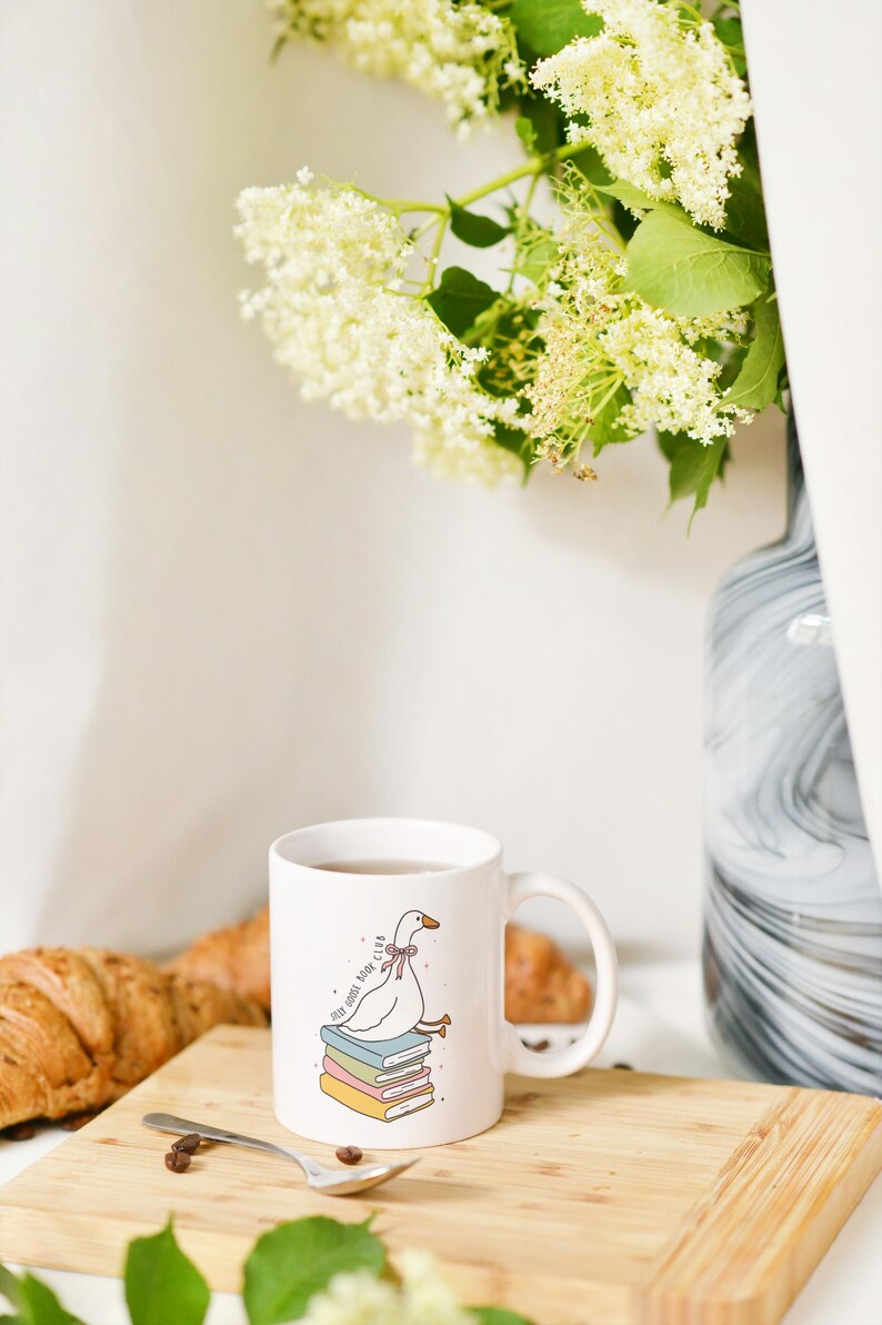 May include: White ceramic mug with a cartoon goose wearing a bow, perched on a stack of books. The mug has the text "Self Goose Book Club". A spoon and coffee beans sit on a wooden cutting board, with a croissant and flowers in the background.