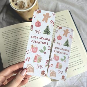 May include: Two rectangular bookmarks with a festive design. The bookmarks feature gingerbread men, Christmas trees, and the text "COSY SEASON ESSENTIALS." The background is a light cream color with a diamond pattern.