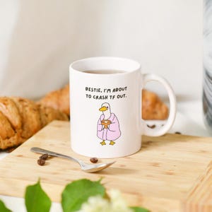May include: White ceramic mug with a cartoon duck wearing a pink robe and holding a cup. The mug has the text "BESTIE, I'M ABOUT TO CRASH TF OUT." on the front. The mug is on a wooden surface with a spoon and coffee beans.