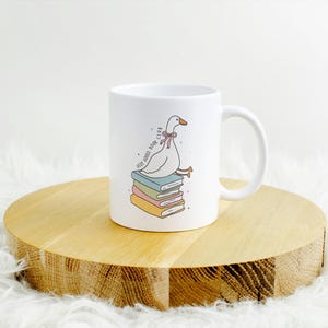 May include: White ceramic mug featuring a cartoon goose with a pink bow tie sitting atop a stack of colorful books. The mug has the text "Very Goose Book Club" and a curved handle. The mug is on a wooden round surface.