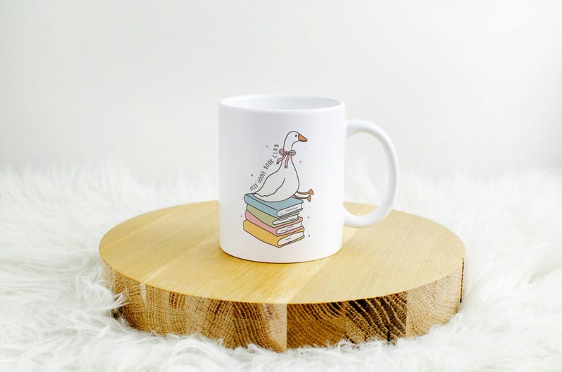 May include: White ceramic mug featuring a cartoon goose with a pink bow tie sitting atop a stack of colorful books. The mug has the text "Very Goose Book Club" and a curved handle. The mug is on a wooden round surface.