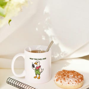 May include: White ceramic mug with the phrase "WHAT THE FUCK, OVER" and a cartoon duck holding a walkie-talkie. The duck wears a red bandana and green overalls. A spoon rests in the mug, which contains tea. A glazed donut and a notebook are also present.
