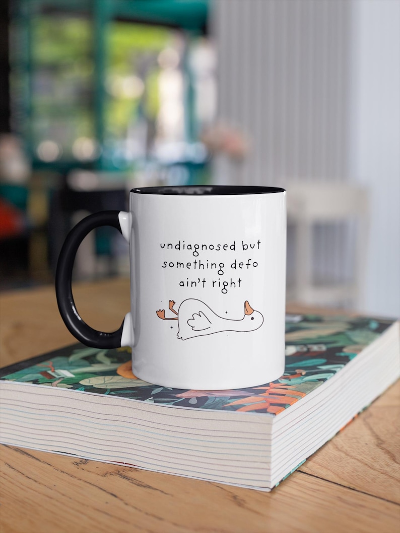 May include: White ceramic mug with a black handle and rim, printed with the text "undiagnosed but something defo ain't right" and a cartoon goose. The mug sits on a book with a patterned cover.