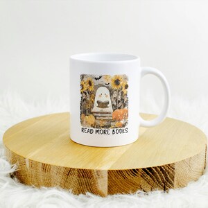 May include: White ceramic mug featuring a whimsical illustration of a ghost reading a book, surrounded by sunflowers and pumpkins. The text "READ MORE BOOKS" is printed below the image. The mug is placed on a wooden surface.