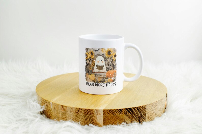 May include: White ceramic mug featuring a whimsical illustration of a ghost reading a book, surrounded by sunflowers and pumpkins. The text "READ MORE BOOKS" is printed below the image. The mug is placed on a wooden surface.