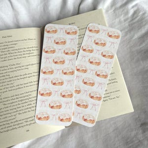 Pode incluir: Dois marcadores retangulares com um padr&atilde;o repetido de rolinhos de canela e la&ccedil;os cor-de-rosa. Os marcadores est&atilde;o colocados nas p&aacute;ginas de um livro aberto. Os marcadores s&atilde;o de cor creme claro.