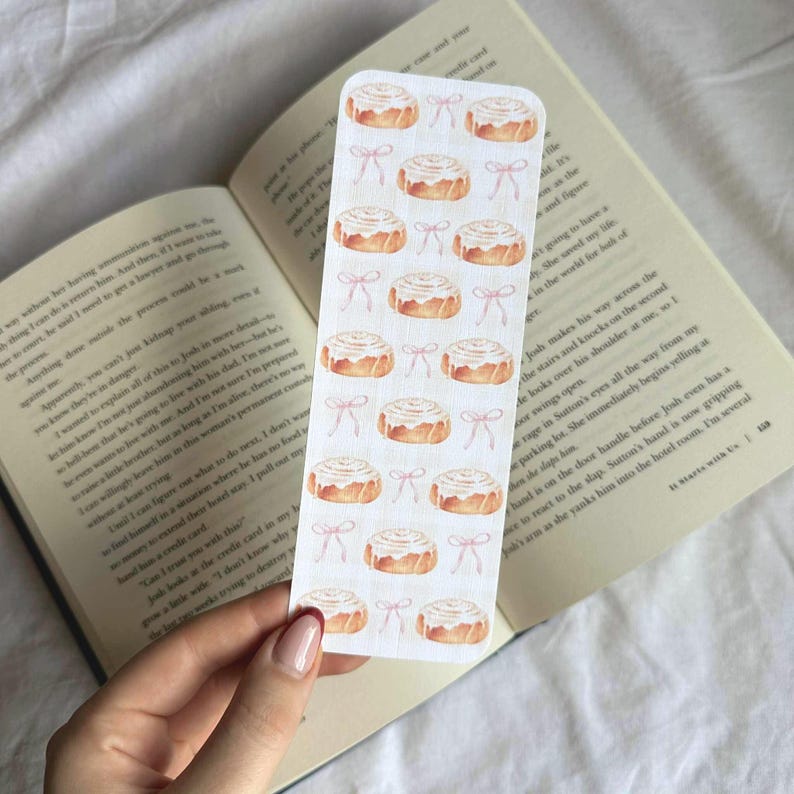 Pode incluir: Um marcador de livros retangular com um padr&atilde;o repetido de rolinhos de canela em aquarela com cobertura branca e la&ccedil;os rosa. O marcador de livros &eacute; segurado sobre um livro aberto.