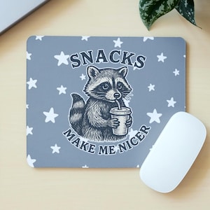 May include: Grey mouse mat featuring a raccoon illustration drinking from a cup. The mat has white stars and the text "SNACKS MAKE ME NICER". A white computer mouse is positioned beside the mat.
