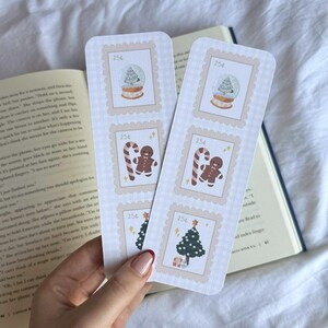 May include: Two festive bookmarks with a postage stamp design. Each bookmark features a snow globe, gingerbread man with a candy cane, and a Christmas tree. The bookmarks are light beige with a checkered pattern.