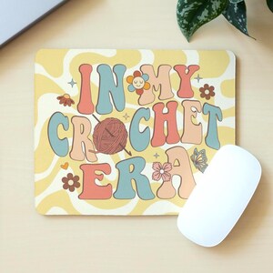 May include: A rectangular mouse mat featuring the text "IN MY CROCHET ERA" in a retro style. The mat has a yellow background with a wavy pattern and decorative flowers and butterflies. A white computer mouse is next to the mat.