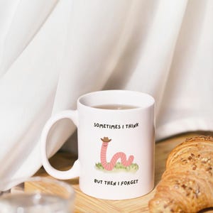 May include: White ceramic mug with a cartoon worm wearing a cowboy hat, the text reads "SOMETIMES I THINK BUT THEN I FORGET". The mug sits on a wooden board with a croissant and a glass of water.