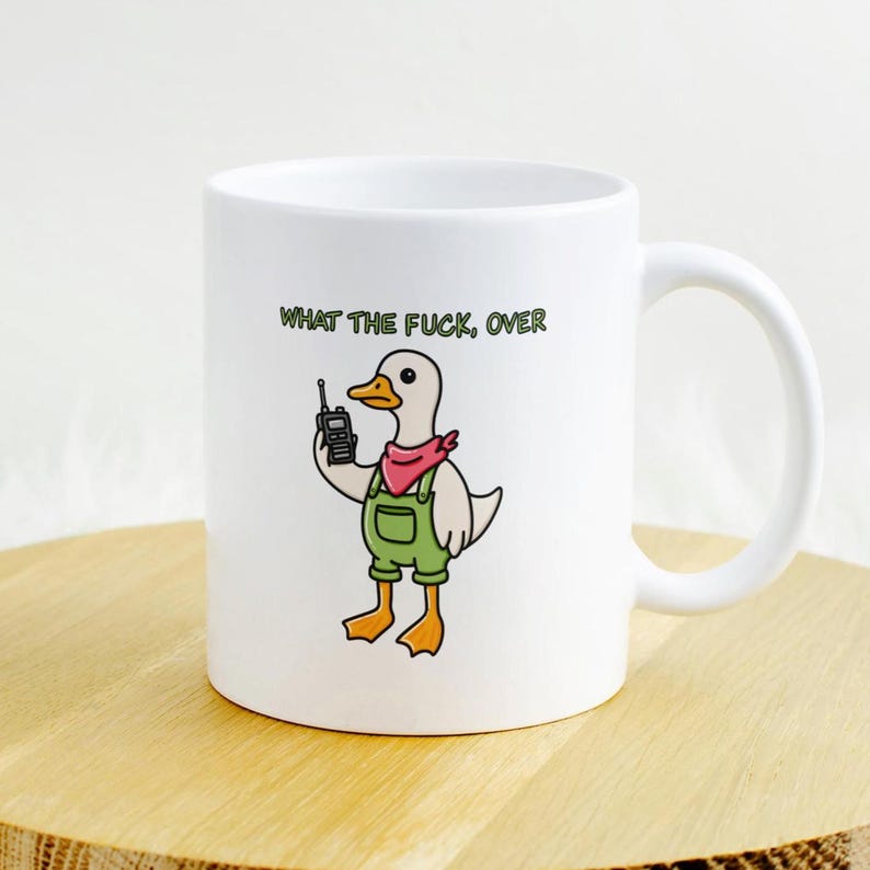 May include: White ceramic mug featuring a cartoon duck wearing green overalls and a red bandana, holding a walkie-talkie. The text "WHAT THE FUCK, OVER" is printed above the duck. The mug has a curved handle.