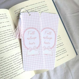 May include: Two pink and white gingham bookmarks with the text "Fell asleep here" in a decorative oval. Each bookmark has a pink tassel and is next to an open book.