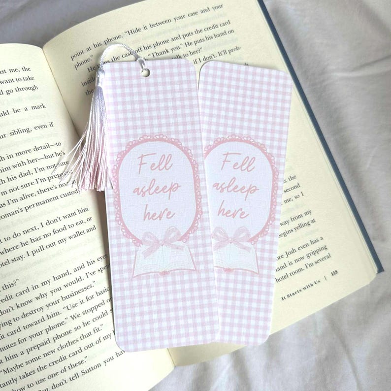 May include: Two pink and white gingham bookmarks with the text "Fell asleep here" in a decorative oval. Each bookmark has a pink tassel and is next to an open book.