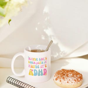 May include: White ceramic mug with the text "MAYBE SHE'S BORN WITH IT MAYBE IT'S ADHD" in colorful retro font. The mug contains tea and a spoon. A glazed donut and a spiral notebook are also on the table.