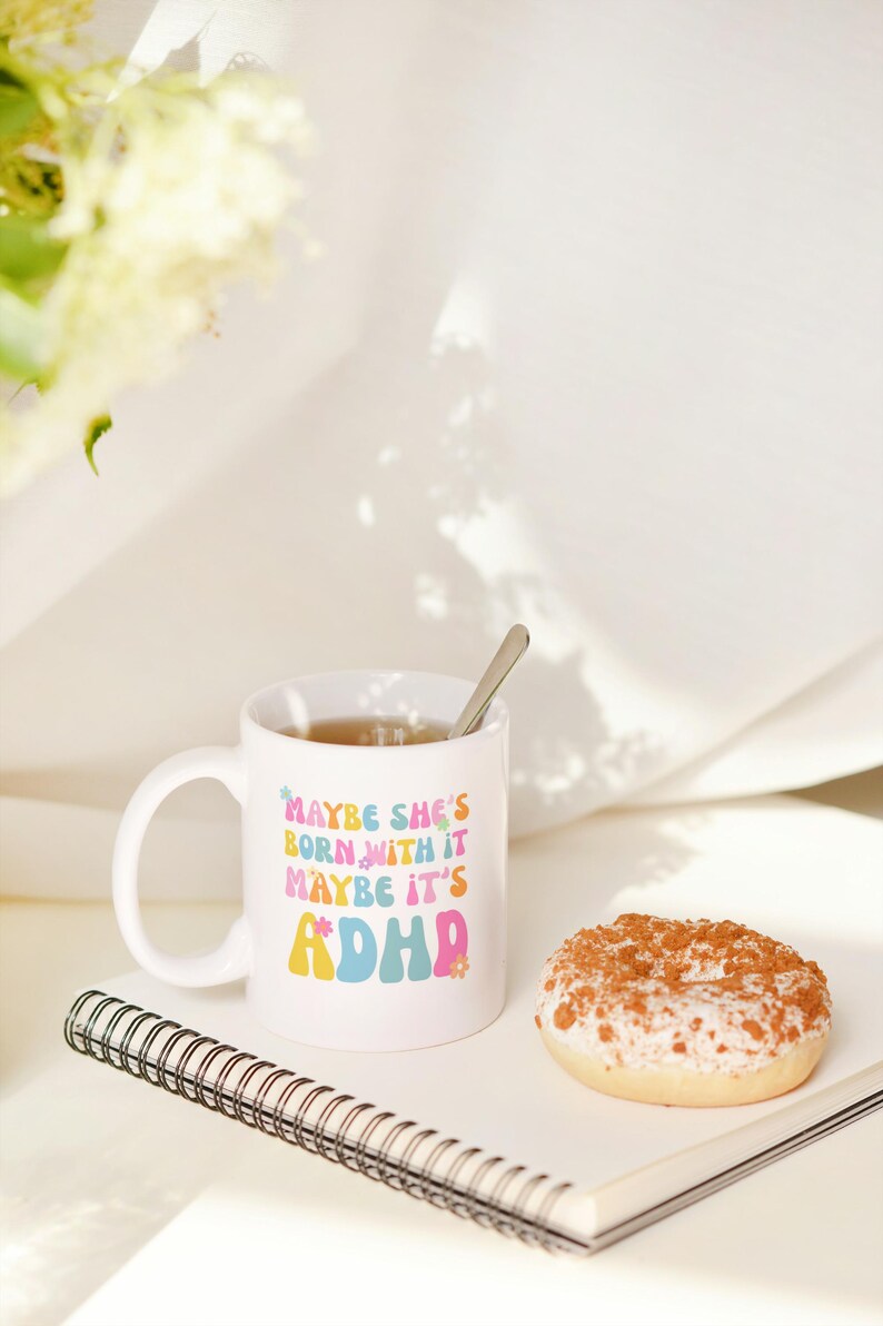 May include: White ceramic mug with the text "MAYBE SHE'S BORN WITH IT MAYBE IT'S ADHD" in colorful retro font. The mug contains tea and a spoon. A glazed donut and a spiral notebook are also on the table.