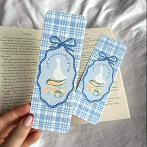 May include: Two blue and white plaid bookmarks with a whimsical design. Each bookmark features a light blue oval with a white goose on books, a teacup, and the text "Just a silly bookish goose."