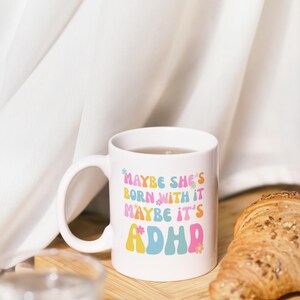 May include: White ceramic mug with the text "MAYBE SHE'S BORN WITH IT MAYBE IT'S ADHD" in colorful retro font. The mug is filled with a clear liquid and sits on a wooden board with a croissant. A glass of water is in the foreground.