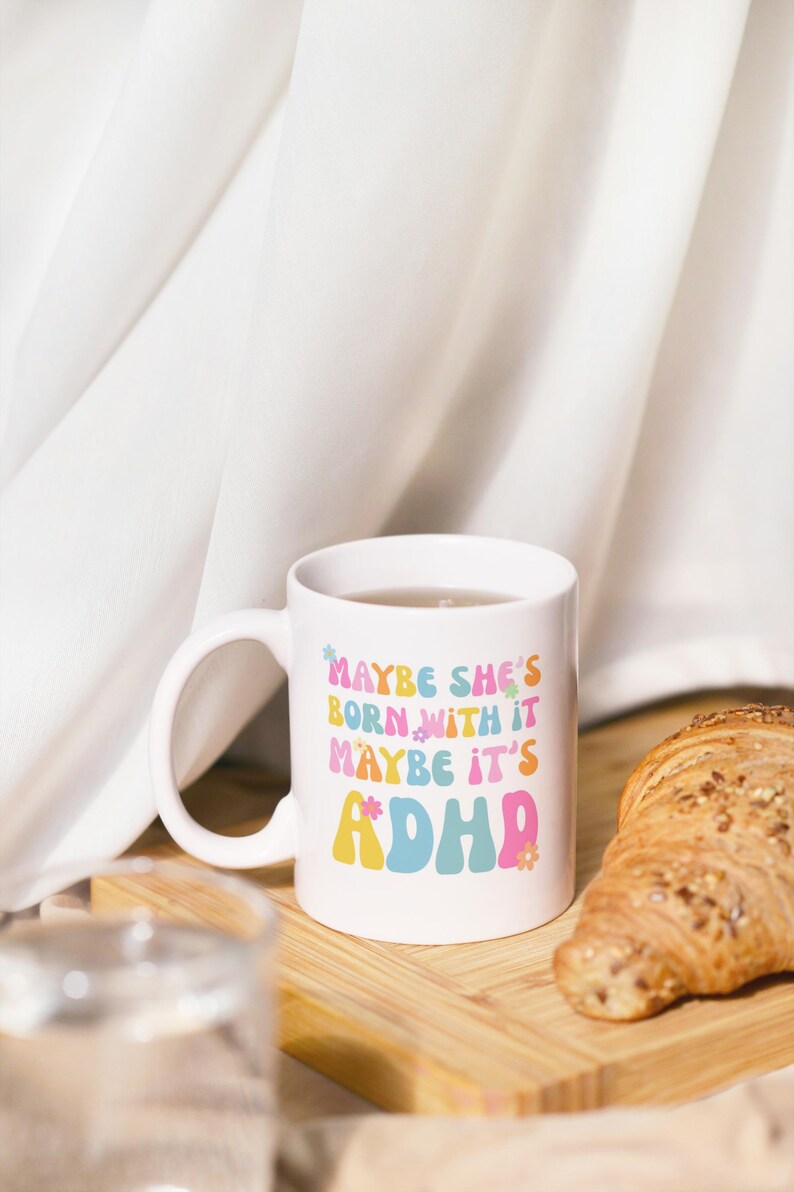 May include: White ceramic mug with the text "MAYBE SHE'S BORN WITH IT MAYBE IT'S ADHD" in colorful retro font. The mug is filled with a clear liquid and sits on a wooden board with a croissant. A glass of water is in the foreground.