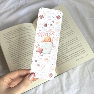 K&ouml;nnte beinhalten: Ein rechteckiger Lesezeichen mit festlichem Design. Es zeigt einen Lebkuchenmann in einer Tasse, die Worte "Hot Chocolate Season" und verschiedene Illustrationen zum Thema Feiertage in sanften Pastellfarben. Das Lesezeichen liegt &uuml;ber einem offenen Buch.