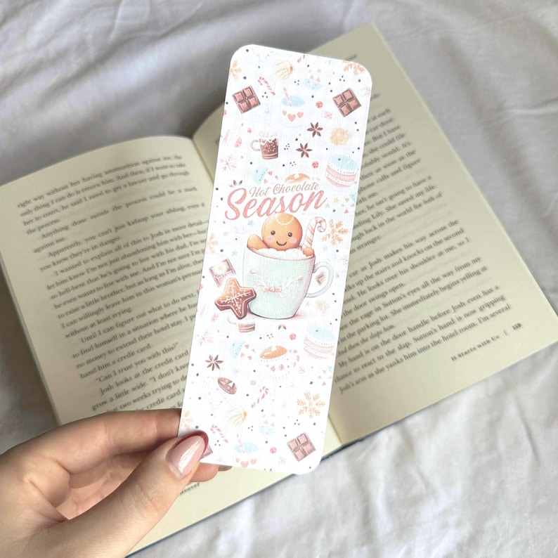 K&ouml;nnte beinhalten: Ein rechteckiger Lesezeichen mit festlichem Design. Es zeigt einen Lebkuchenmann in einer Tasse, die Worte "Hot Chocolate Season" und verschiedene Illustrationen zum Thema Feiertage in sanften Pastellfarben. Das Lesezeichen liegt &uuml;ber einem offenen Buch.