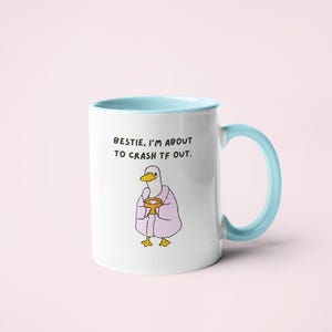 May include: White ceramic mug with a light blue interior and handle. The mug features a cartoon duck wearing a pink robe and holding a cup. Text on the mug reads: "Bestie, I'm about to crash tf out."