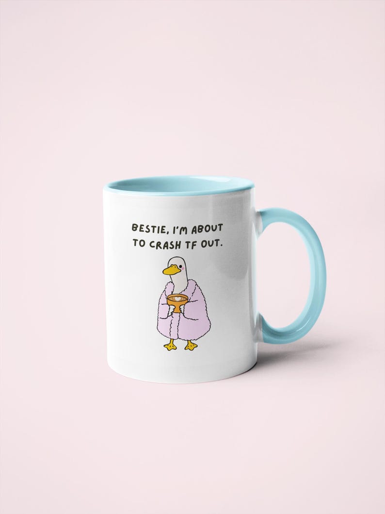 May include: White ceramic mug with a light blue interior and handle. The mug features a cartoon duck wearing a pink robe and holding a cup. Text on the mug reads: "Bestie, I'm about to crash tf out."