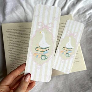 May include: Two decorative bookmarks with a whimsical design. Each bookmark features a white goose atop a stack of books and a teacup, framed by a pink border and bows. The background has vertical stripes in light green and white.