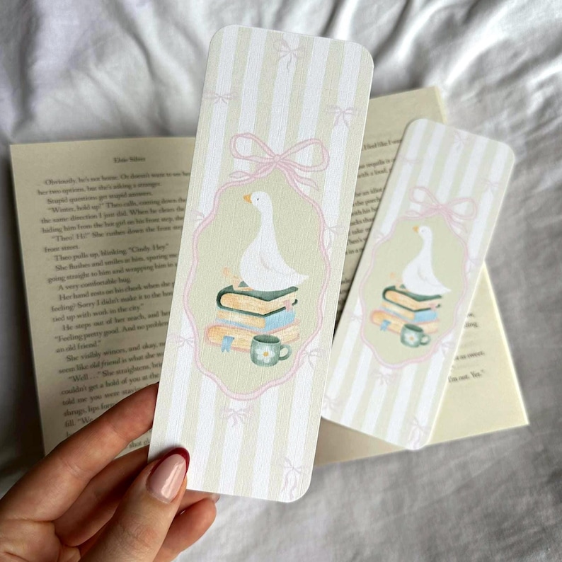 May include: Two decorative bookmarks with a whimsical design. Each bookmark features a white goose atop a stack of books and a teacup, framed by a pink border and bows. The background has vertical stripes in light green and white.