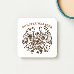 May include: Square coaster featuring a cartoon illustration of two frogs in sweaters, each holding a mushroom. A basket of mushrooms is between them. The words "SWEATER WEATHER" are printed above in brown.