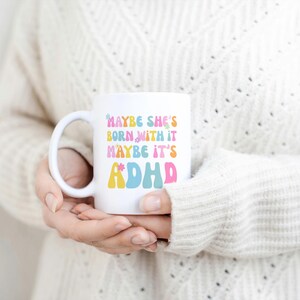 May include: White ceramic mug with the text "MAYBE SHE'S BORN WITH IT MAYBE IT'S ADHD" in colorful, retro-style lettering. The mug has a curved handle and is held by a person wearing a cream-colored sweater.