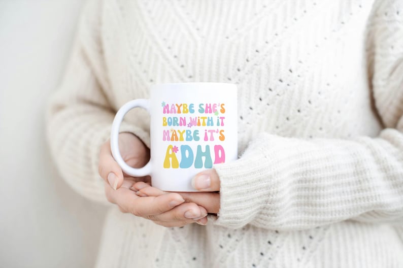 May include: White ceramic mug with the text "MAYBE SHE'S BORN WITH IT MAYBE IT'S ADHD" in colorful, retro-style lettering. The mug has a curved handle and is held by a person wearing a cream-colored sweater.