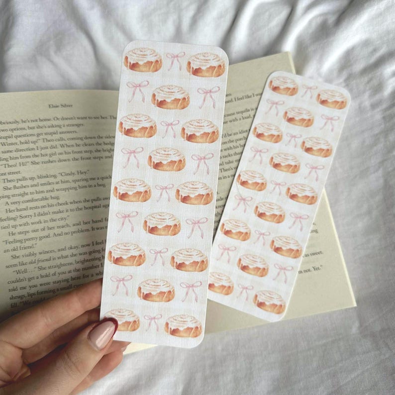 Pode incluir: Dois marcadores de livros de papel com um padr&atilde;o repetido de rolinhos de canela e la&ccedil;os cor-de-rosa. Os marcadores s&atilde;o retangulares. Um livro com texto &eacute; vis&iacute;vel no fundo.
