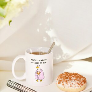 May include: White ceramic mug with the text "Bestie, I'm about to crash TF out." and a cartoon duck wearing a pink robe. A donut with white frosting and brown sprinkles sits next to the mug on a notebook.