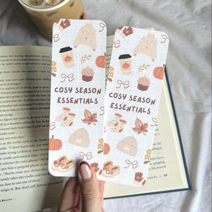 May include: Two rectangular bookmarks with a grid pattern, featuring fall-themed illustrations like pumpkins, coffee cups, and sweaters. The text "COSY SEASON ESSENTIALS" is printed on each bookmark.