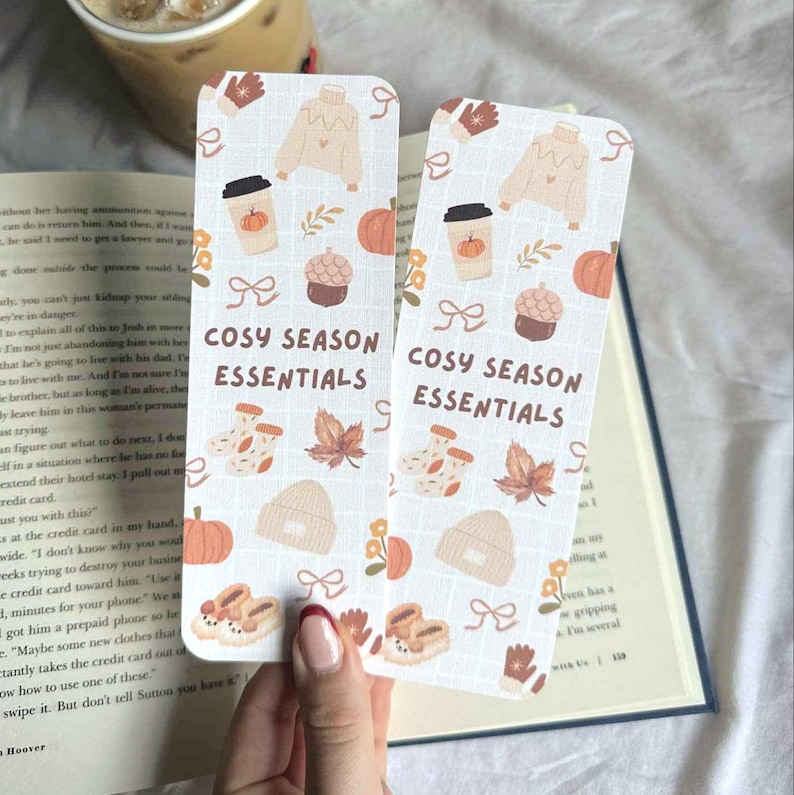 May include: Two rectangular bookmarks with a grid pattern, featuring fall-themed illustrations like pumpkins, coffee cups, and sweaters. The text "COSY SEASON ESSENTIALS" is printed on each bookmark.