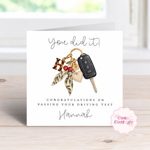 May include: A white greeting card featuring the text You did it! and Congratulations on passing your driving test Hannah. The illustration shows a gold keyring with a black car key, a leopard print letter H, two red cherries, and a heart charm.
