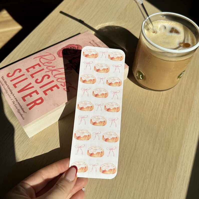 Pode incluir: Um marcador de livros com ilustra&ccedil;&otilde;es de rolinhos de canela e la&ccedil;os cor-de-rosa. O marcador est&aacute; perto de um livro intitulado "Reckless" de Elsie Silver e um copo de caf&eacute; numa superf&iacute;cie de madeira.