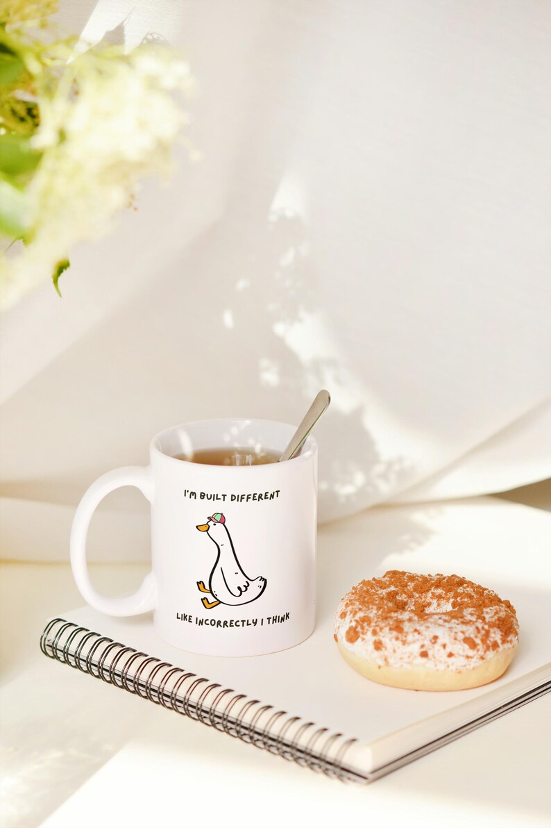 Puede incluir: Taza de cer&aacute;mica blanca con una ilustraci&oacute;n de un ganso de dibujos animados y el texto "I'm built different like incorrectly I think". Una cuchara descansa en la taza, que contiene t&eacute;. Tambi&eacute;n hay una rosquilla glaseada y un cuaderno espiral.
