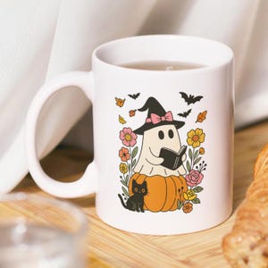 May include: White ceramic mug with a Halloween design. A ghost in a witch's hat reads a book while sitting on an orange pumpkin, surrounded by flowers, leaves, bats, and a black cat. The mug has a curved handle.