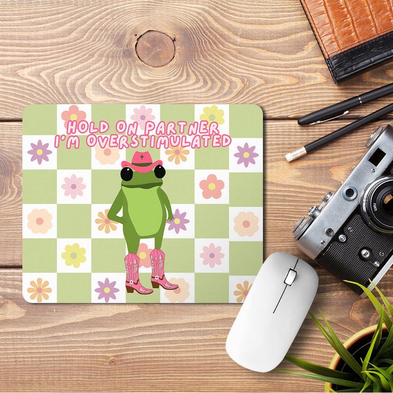 Op de afbeelding: Een groene kikker met een roze cowboyhoed en roze laarzen op een groen-wit geruite achtergrond met roze bloemen. De tekst "HOLD ON PARTNER I'M OVERSTIMULATED" staat in zwarte letters.