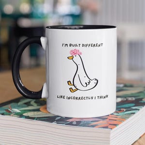 Könnte beinhalten: Weiße Keramik-Tasse mit schwarzem Henkel und Rand. Die Tasse zeigt eine Cartoon-Ente mit einer rosa Blume und dem Text "I'M BUILT DIFFERENT LIKE INCORRECTLY I THINK". Die Tasse steht auf einer gemusterten Oberfläche.