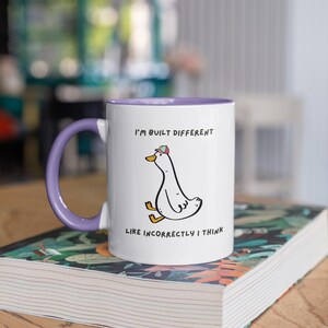 Puede incluir: Taza de cer&aacute;mica blanca con interior y asa morados. La taza presenta un pato de dibujos animados con sombrero y el texto "I'M BUILT DIFFERENT LIKE INCORRECTLY I THINK". La taza est&aacute; sobre un libro.