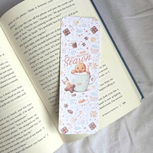 K&ouml;nnte beinhalten: Dekorativer Lesezeichen mit dem Motiv "Hot Chocolate Season". Es zeigt einen Lebkuchenmann in einer Tasse, umgeben von S&uuml;&szlig;igkeiten und Schneeflocken. Der Text ist in einer verspielten Schriftart.