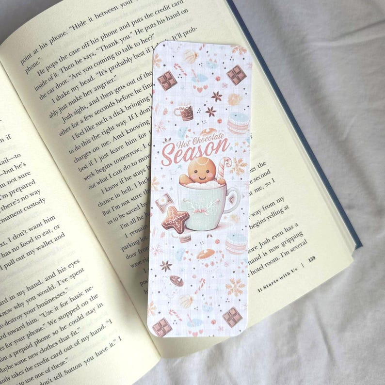 K&ouml;nnte beinhalten: Dekorativer Lesezeichen mit dem Motiv "Hot Chocolate Season". Es zeigt einen Lebkuchenmann in einer Tasse, umgeben von S&uuml;&szlig;igkeiten und Schneeflocken. Der Text ist in einer verspielten Schriftart.