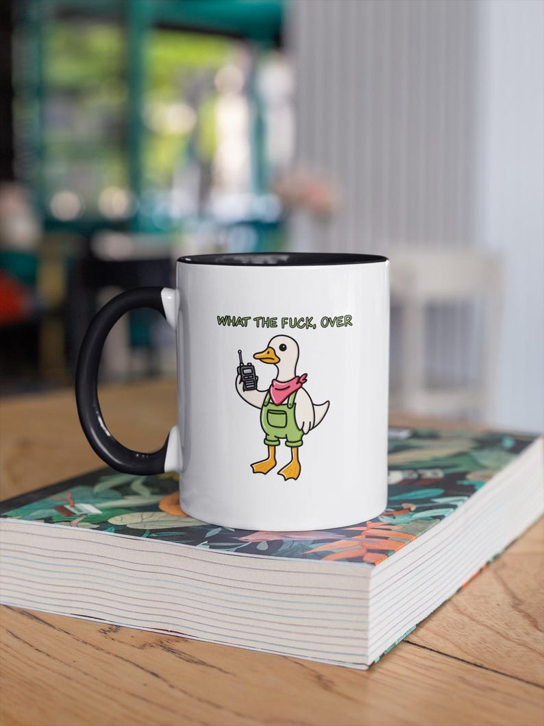 May include: White ceramic mug with a black handle and interior. The mug features a cartoon duck wearing overalls and a pink bandana, holding a cell phone. The text "WHAT THE FUCK, OVER" is printed above the duck.